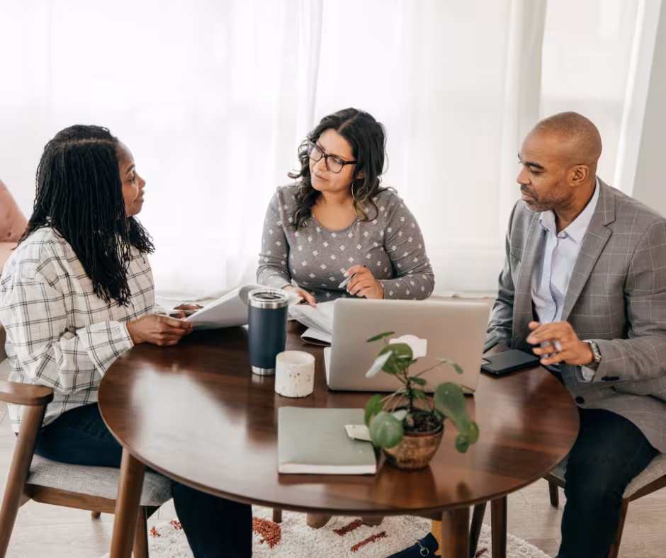 Three people in discussion sat around a round table, with a laptop and notebooks on the table